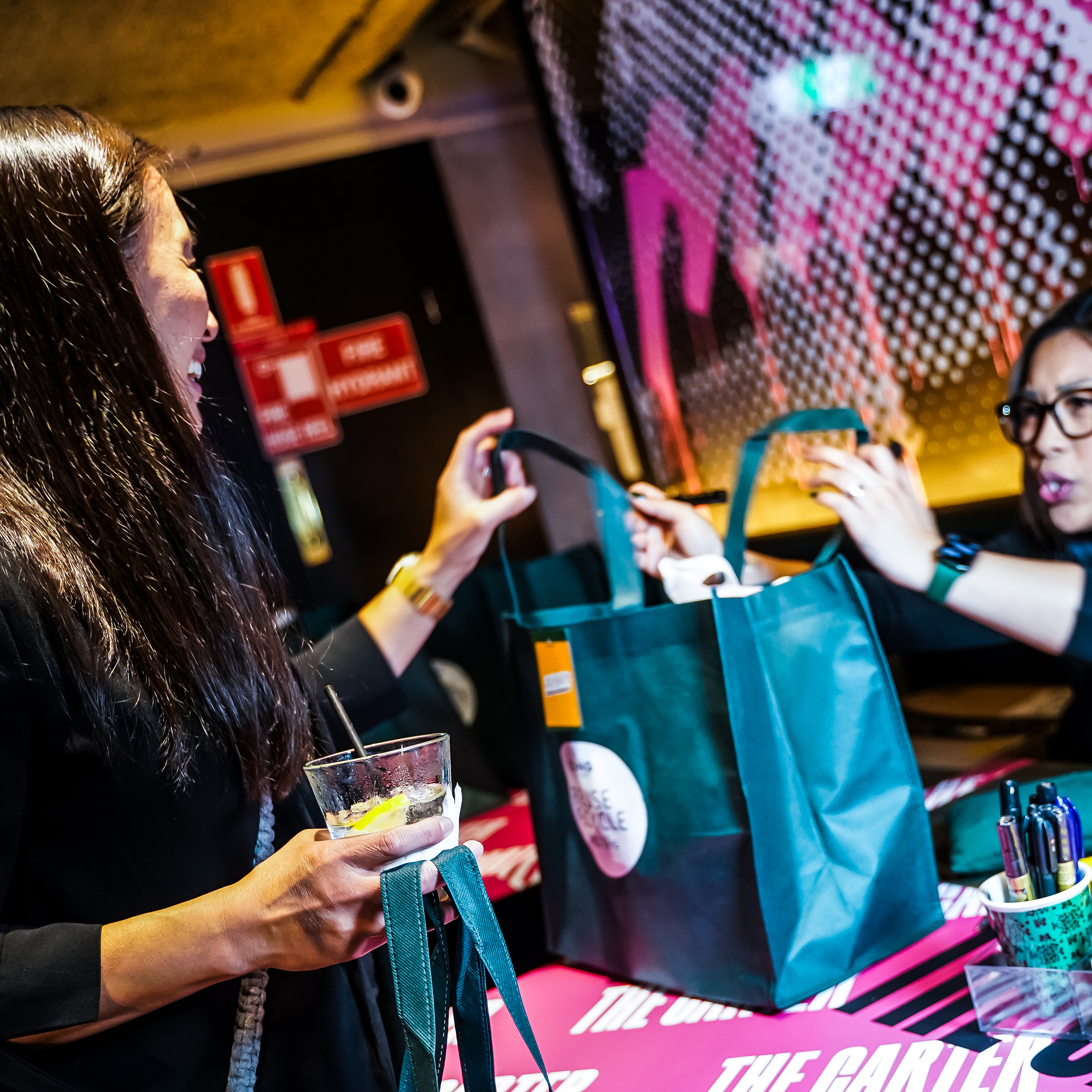 A guest at a brand activation event interacting with a calligrapher while receiving a personalized fabric bag. The setting features a vibrant atmosphere with branded decor and tools for customisation