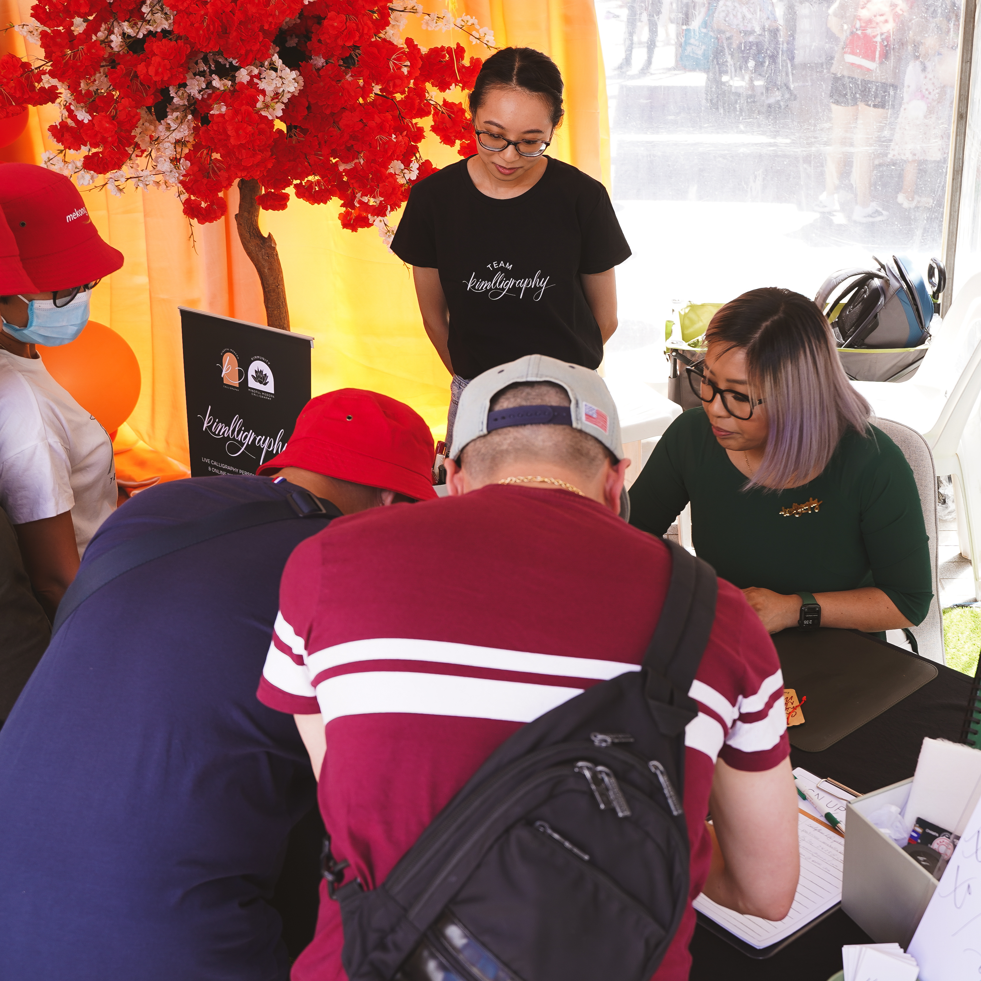 Kim, founder of Kimlligraphy, working on custom hand-lettered designs on wood at an outdoor event. Attendees observe the process while wooden pieces and calligraphy tools are displayed on the table.